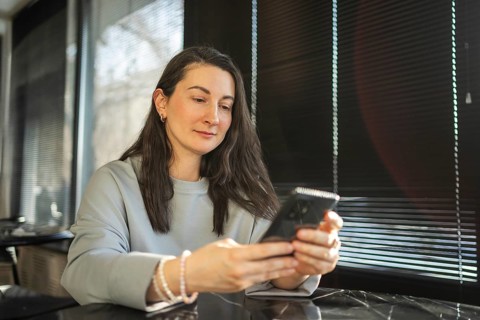 A mid-aged woman is casually using her smartphone in a sunlit cafe environment, reflecting a moment