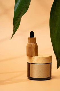 A cosmetic bottle with a wooden cap on a warm beige background, accented by green leaves. The