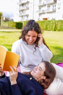 A young women smiles while lying down looking at her partner, who is reading from an orange book in