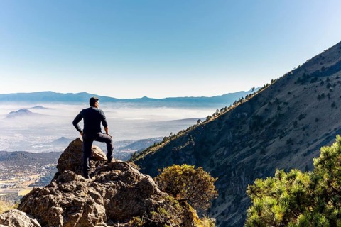 A person stands triumphantly on a rocky mountain peak, gazing over a vast, sunlit valley with