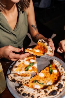 A person holds a slice of fresh Margherita pizza topped with mozzarella and basil. The pizza is