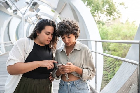 A lesbian couple shares a moment on a contemporary walkway, looking at a smartphone. The scene