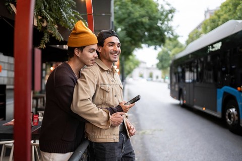 A joyful gay couple embraces by a bus stop. They are smiling, holding a phone, with trees,