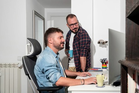 Two gay men collaborate in a cozy home office, mixing work with casual interaction. One sits at a