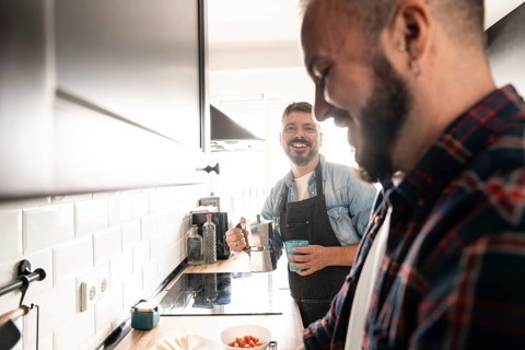 Two gay men enjoy preparing a meal together in a modern kitchen. One holds a coffee pot while the
