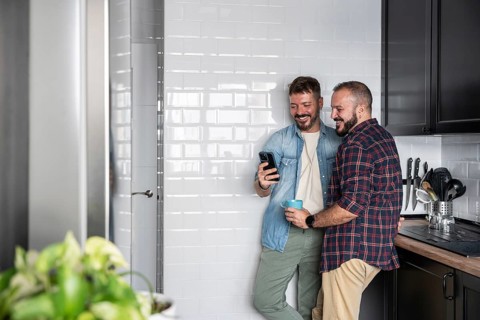 A happy gay couple stands in a bright kitchen, sharing a joyful moment while looking at a