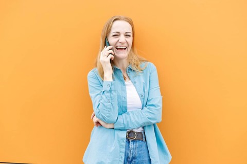 A cheerful woman in a blue shirt talks on her smartphone against an orange background in Warsaw,