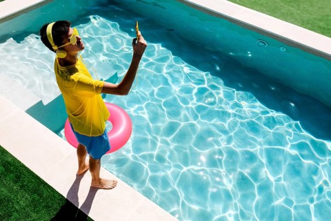 A young teenage boy in vibrant yellow attire takes a selfie by a sparkling blue pool. Sunlight