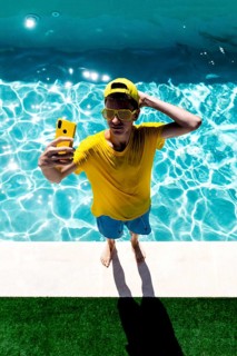 A teenage boy in a yellow shirt, sunglasses, and cap takes a selfie by a bright blue pool. His