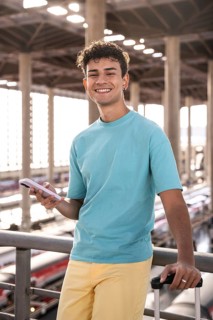A cheerful young man stands at a train station, holding a phone and a suitcase. He is smiling and