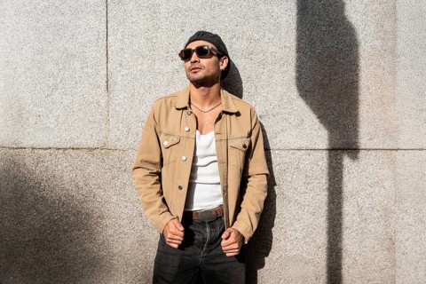 A man in a tan jacket and sunglasses leans against a sunlit concrete wall. The casual attire and