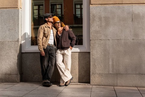 A Colombian gay couple in casual outfits enjoys a sunny urban setting. They lean against a