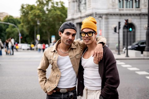 A Colombian gay couple stands joyfully in an urban setting. They wear casual outfits, expressing a