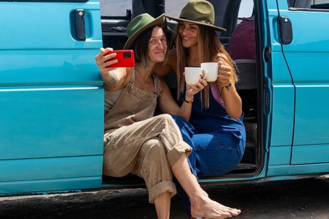 Two lesbians women enjoying a road trip sit by a turquoise van. They smile, capturing the moment