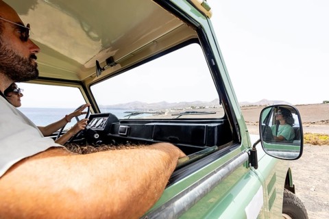 A blind man and a woman drive a motorhome along a coastal road with scenic mountains in the