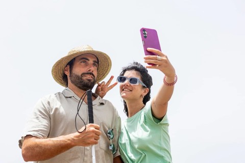 A blind man and a woman smile while taking a playful selfie by the sea. The woman makes a peace
