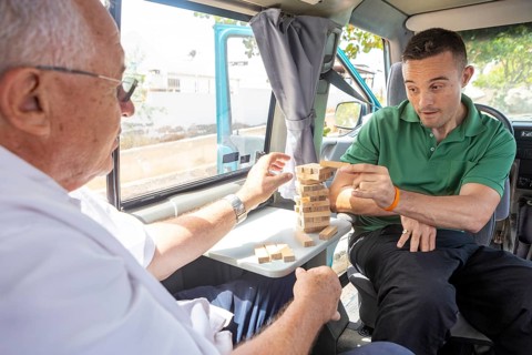 An elderly man and his adult son enjoy playing a game of Jenga inside a van, parked in a sunny