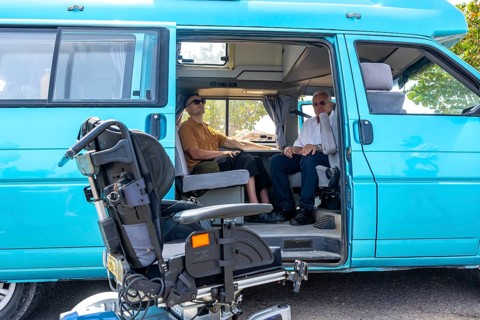 Two men sitting inside a blue van that's adapted for wheelchair access, with the wheelchair