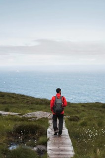 A traveler wearing a red jacket and backpack walks along a wooden path overlooking the Norwegian