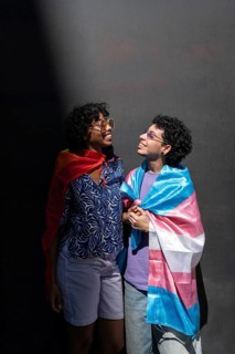 A joyful multiethnic LGBTQ+ couple smiling at each other, wrapped in Pride flags. Captured in