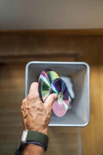A person hand dropping assorted DVDs into a trash bin. The image highlights the decline of physical