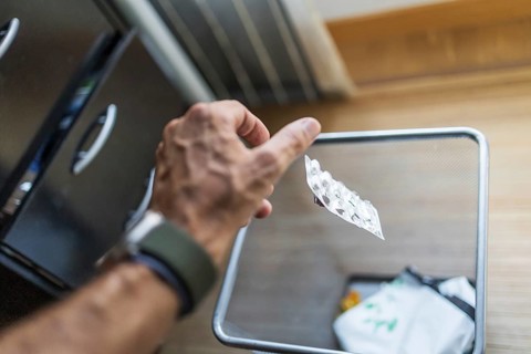 A hand disposes of an blister pack into a mesh waste bin. The casual scene highlights medication
