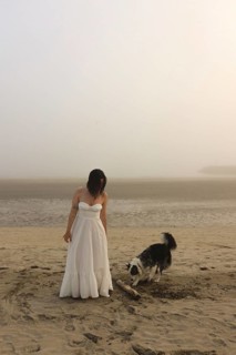 A woman in a white dress stands on a misty beach with a playful dog nearby The serene atmosphere