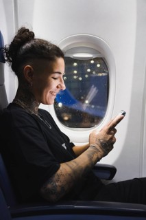 A young woman with intricate tattoos sits on an airplane, smiling while using a smartphone The