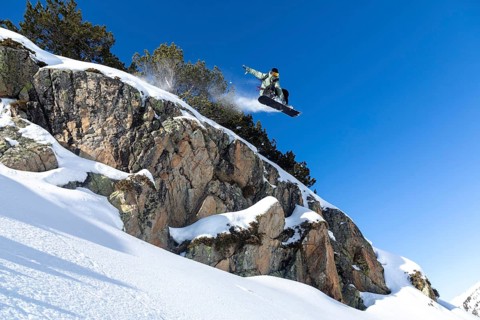 A snowboarder catches air while leaping off a rocky cliff, against a vibrant blue sky. The scene