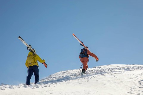 Two skiers in colorful gear hike up a snowy slope, carrying their skis The clear blue sky contrasts