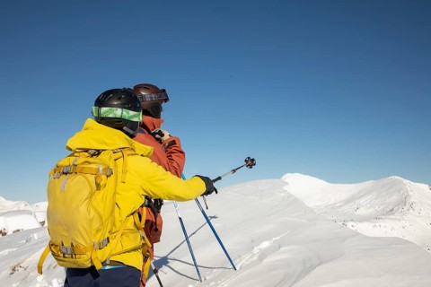 Two freeride skiers stand atop a snow-covered peak, gazing into the vast mountain range under a