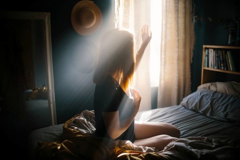 A woman sits on a bed, bathed in morning sunlight streaming through the window. The warm glow