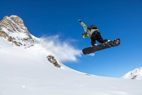 A snowboarder performs a jump against a clear blue sky, with snow spraying behind. The stunning