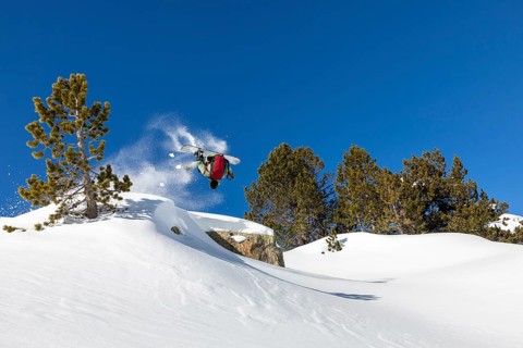A snowboarder performs a backflip against a clear blue sky in a snow covered mountain landscape.