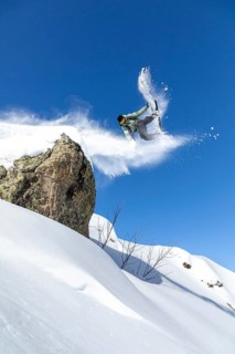 A snowboarder performs an aerial trick off a snowy cliff against a clear blue sky, capturing the