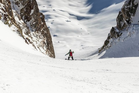 A snowboarder carves down a steep, snow covered slope between rocky peaks. The bright snow