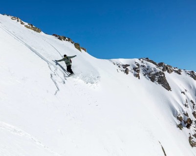 A snowboarder skillfully descends a steep, snowy slope against a clear blue sky. The untouched snow