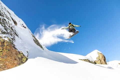 A snowboarder executes an impressive jump, sending a spray of snow flying. The scene is set against