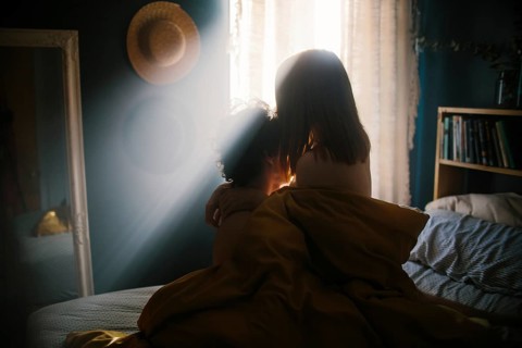 A couple shares an intimate embrace on a bed, bathed in warm morning light streaming through a