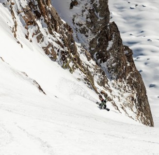 A snowboarder skillfully descends a steep, snowy slope surrounded by rugged cliffs and untouched