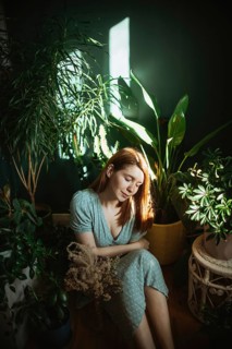 A woman in a light dress calmly sits surrounded by lush indoor plants. Sunlight filters through,