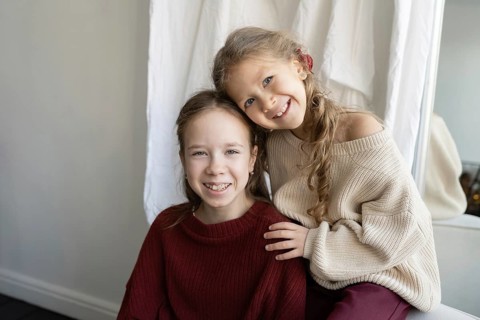 Two sisters wearing cozy sweaters smile warmly at the camera The younger sibling leans