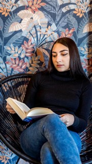 A young woman is engrossed in reading a book while sitting in a modern wicker chair, against a