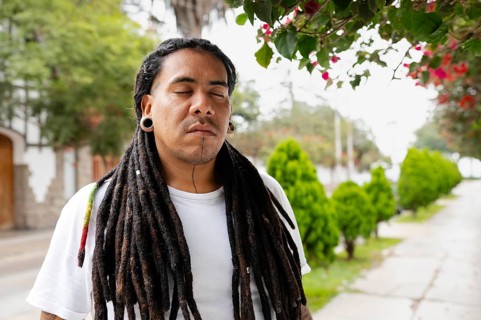 A man with long dreadlocks stands peacefully with his eyes closed under blooming bougainvillea