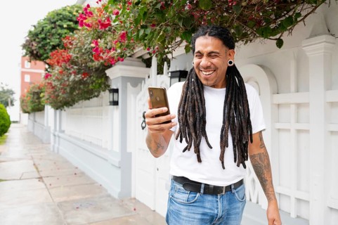 A joyful man with dreadlocks holds a smartphone, smiling widely as he stands in front of a vibrant,
