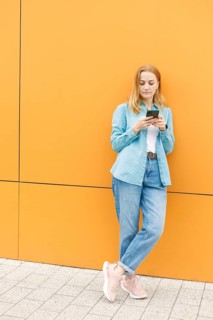 A woman leans against a bright orange wall in Warsaw, focused on her phone She wears a blue shirt,