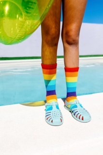 Vibrant scene featuring legs in rainbow socks and teal sandals, beside a pool. The bright turquoise