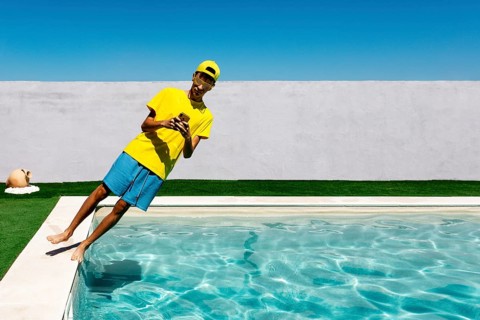 A teenage boy in vibrant yellow attire leans playfully by a pool, capturing a moment with a phone.