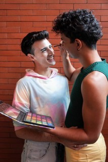 A couple shares a tender moment as one applies makeup to the other with a colorful palette, set