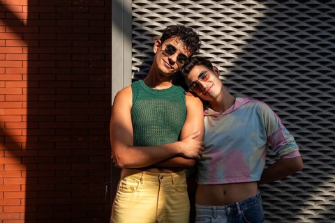 A joyful gay couple in stylish outfits stands together, one leaning on the other affectionately.